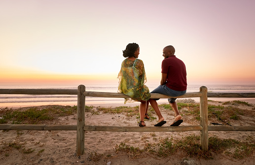 Rear view of beautiful young couple sitting on fence along the beach and talking