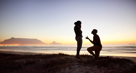 Silhouette of young man staying on the knee and making proposal with an engagement ring for his girl on the beach with evening sunlight on background at the beach