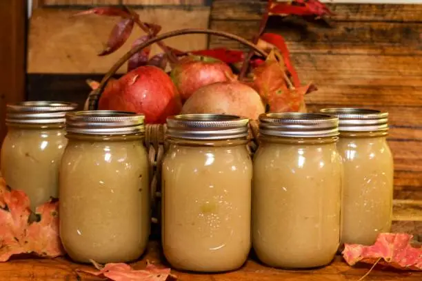 Glass jars of homemade applesauce with basket of apples and autumn leaves