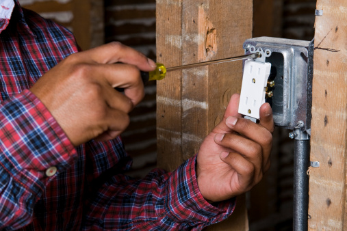 Portrait of a tan young adult latino male construction worker wearing a plaid red and blue long sleeve shirt screwing in an electrical outlet into the metal mainframe.