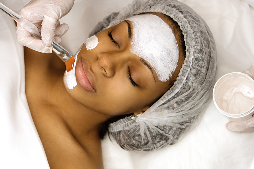Application of a cosmetology mask on the face of a young afro american woman. Procedure for face skin rejuvenation. Beauty, spa, cosmetology and preservation of youth. Wellness relaxation concept.