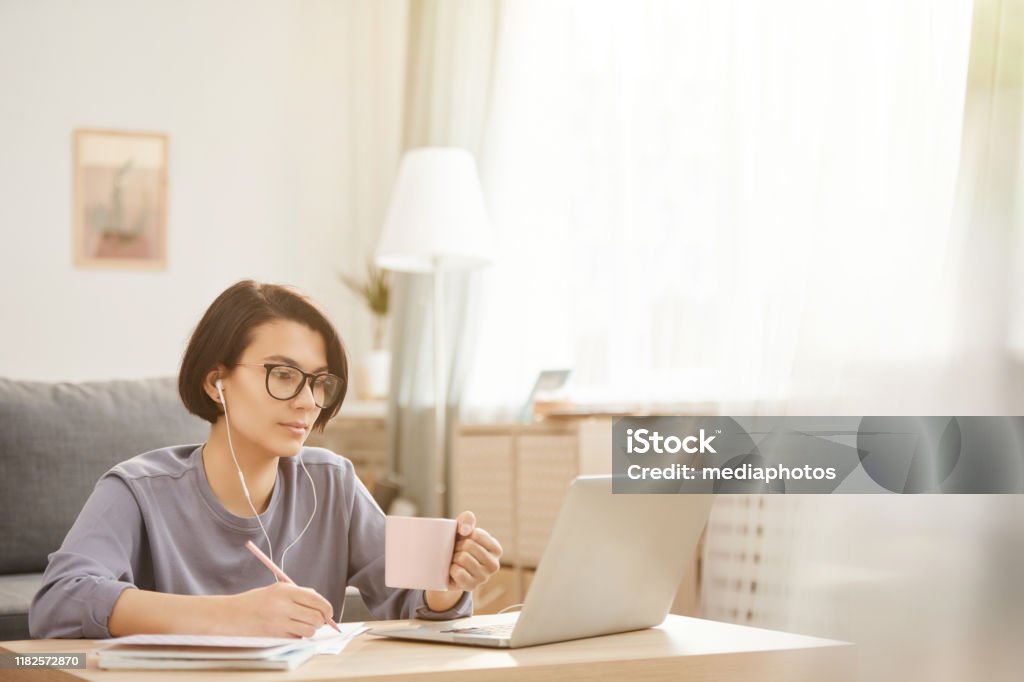 Serious thoughtful young freelance specialist in glasses sitting at coffee table in cozy apartment and drinking coffee while transcribing audio to text Transcribing audio to text E-Learning Stock Photo