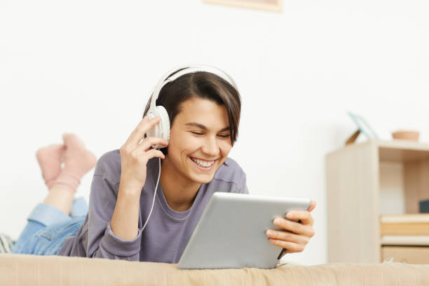 istock Cheerful attractive girl in wired headphones lying on bed at home and using tablet while having in-person conversation via technology 1182572984