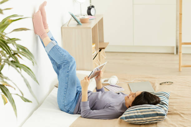 istock Cheerful attractive young woman in jeans lying on bed and leaning feet on wall while using tablet and spending time on social media apps 1182573037