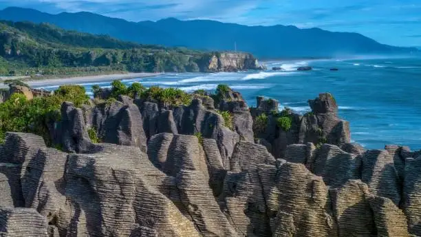 In the foreground is a large set of unusual pinnacle rock formations on land, with grooves eroded into the limestone by water coming through a blowhole, making them look like pancakes. In the background is a beautiful seascape, with a beach, cliff, and low mountain range, while waves from the Tasman Sea break along the shoreline.