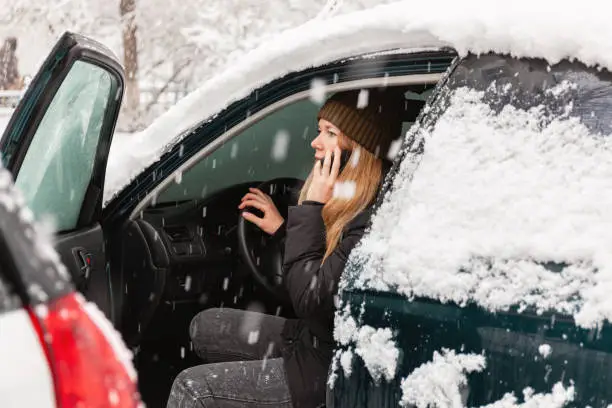 Photo of Young woman calling for help or assistance inside snow covered car. Engine start in frost. Breakdown services in the winter