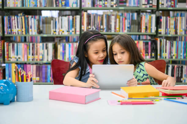 due piccole ragazze carine felici che giocano su un dispositivo di elaborazione tablet pc in biblioteca a scuola. concetto di tecnologia wireless per l'istruzione e l'autoapprogo. stili di vita e amicizia delle persone. bambini in età prescolare - humor book fun human age foto e immagini stock