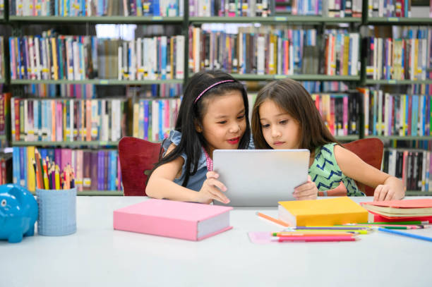 две маленькие счастливые милые девочки, играющие на планшете пк вычислительного устройства в библиотеке в школе. образование и самостояте� - humor book fun human age стоковые фото и изображения
