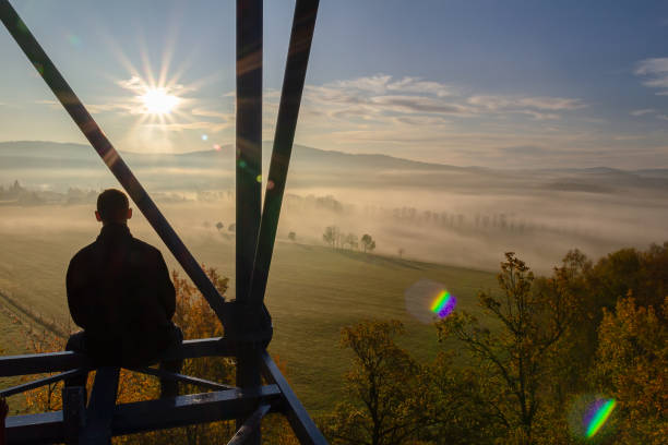 istock Young man sitting from back on watchtower construction looking to autumn foliage trees with misty fog and hill at sunrise, Czech landscape 1182613084