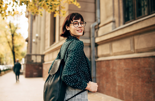 Rear view outdoor image of beautiful young woman with backpack going to college. Young female university student walking beside building campus in the city street.