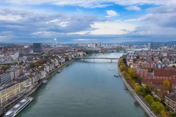 An aerial view of the Basel Switzerland above the Rhine River. The city is waking up with  a dramatic sky. Autumn colors along the shore. Boats are visible in the Rhine River.