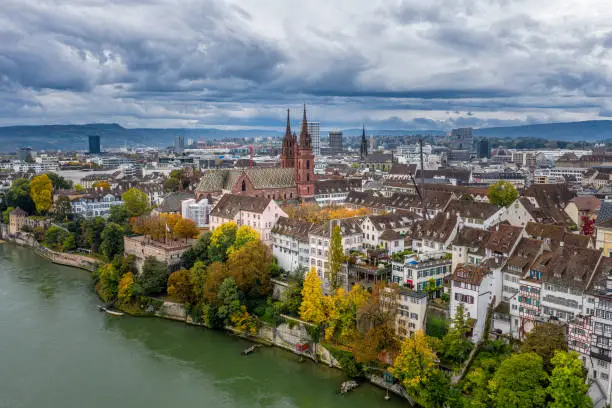 An aerial view of the Basel Switzerland above the Rhine River. A view of Basler Munster Cathedral and it's tall spires in the morning light. A dramatic sky and bright Autumn colors along the shore.