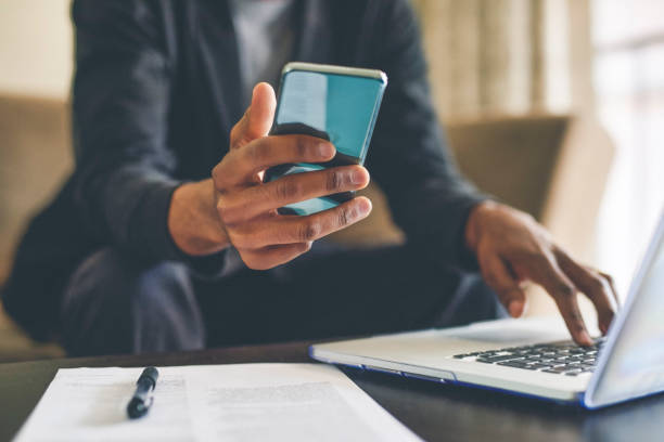 Connecting from the comfort of his home Cropped shot of an unrecognizable man using a smartphone and a laptop while working from home portable information device stock pictures, royalty-free photos & images