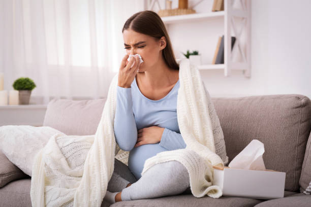 mujer embarazada soplando nariz en tejido sentado en el sofá interior - sneezing tissue young adult cold fotografías e imágenes de stock