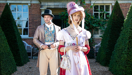 Handsome man and beautiful woman dressed in vintage clothing, standing in front of stately brick home