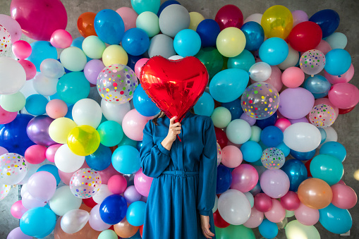 In front of wall with multi colored balloons standing woman in dress and holding heart shaped helium balloon, covering face
