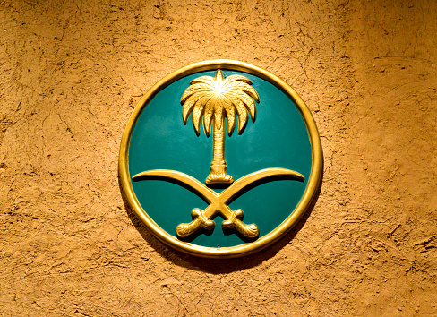 Riyadh, Kingdom of Saudi Arabia:  Saudi Arabian national emblem - two crossed swords with a palm tree in the space above and between the blades - the palm stands for prosperity and the crossed swords represent justice - Saudi coat of arms - placed on a mud covered wall.