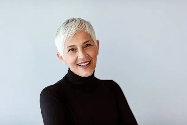 Casual portrait of happy mature woman with natural white short hair and minimal makeup. standing against gray wall.