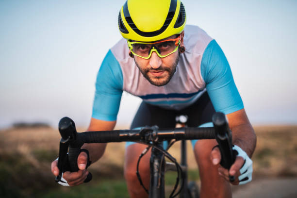 An athlete is riding a bicycle on track. The man is wearing black bike shorts and shin guards along with a red sleeveless top and a red and white helmet and sunglasses. The image is blurred in motion.