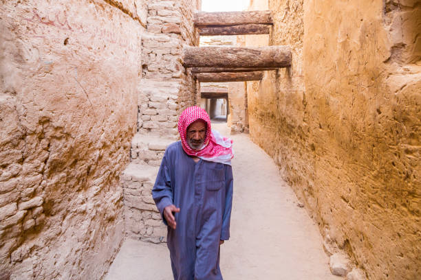 old arabian egyptian man walking dressed in blue national male dress thawb, medieval street through ancient islamic town in al qasr built of mud bricks. motion - desert egyptian culture village town imagens e fotografias de stock