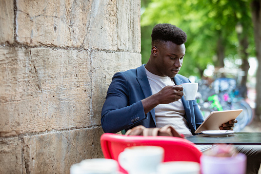 Businessman sitting down at an outdoor table in a city while drinking a hot drink and looking at his digital tablet.