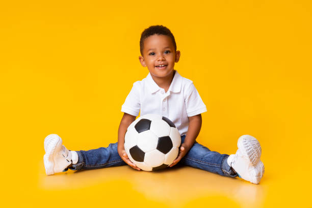 porträt von niedlichen kleinen jungen mit fußball sitzen über gelben studio hintergrund - soccer child indoors little boys stock-fotos und bilder