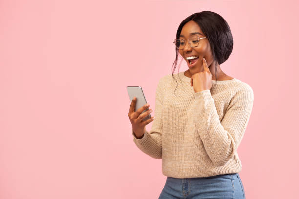 возбужденная девушка с помощью смартфона держа палец на подбородке, розовый фон - tone dialing стоковые фото и изображения