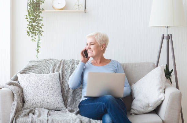 istock Modern old lady holding laptop and talking by phone 1182647558