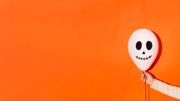 Creepy white balloon with ghost face in woman hand on bright orange background