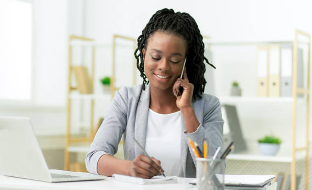 istock Smiling Woman Talking On Cellphone Taking Notes Sitting At Workplace 1182647703