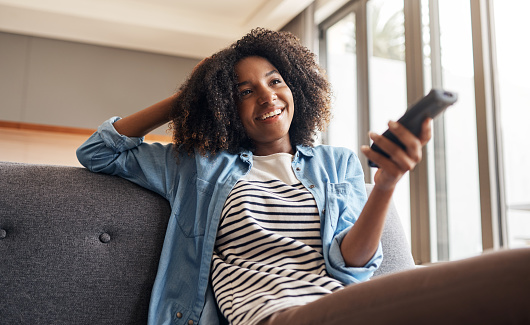 Shot of a young woman holding a remote control while sitting on the sofa at home