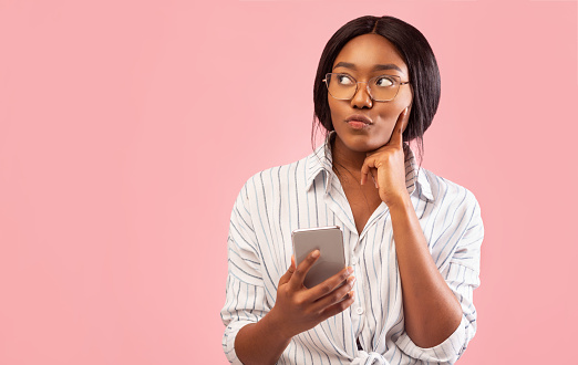 Thoughtful African American Girl Using Smartphone Thinking About Something Standing Over Pink Studio Background. Free Space