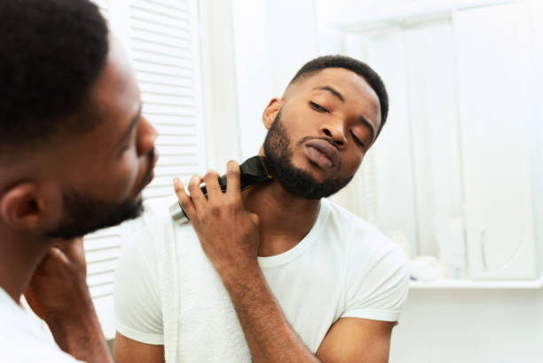 Young black man looking at mirror and shaving beard with trimmer Grooming concept. Young black man looking at mirror and shaving beard with trimmer or electric shaver at bathroom, free space beard stock pictures, royalty-free photos & images