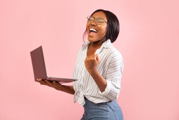 emocionada afro chica sosteniendo portátil gesturing sí, tiro de estudio - vertical studio shot indoors pink fotografías e imágenes de stock
