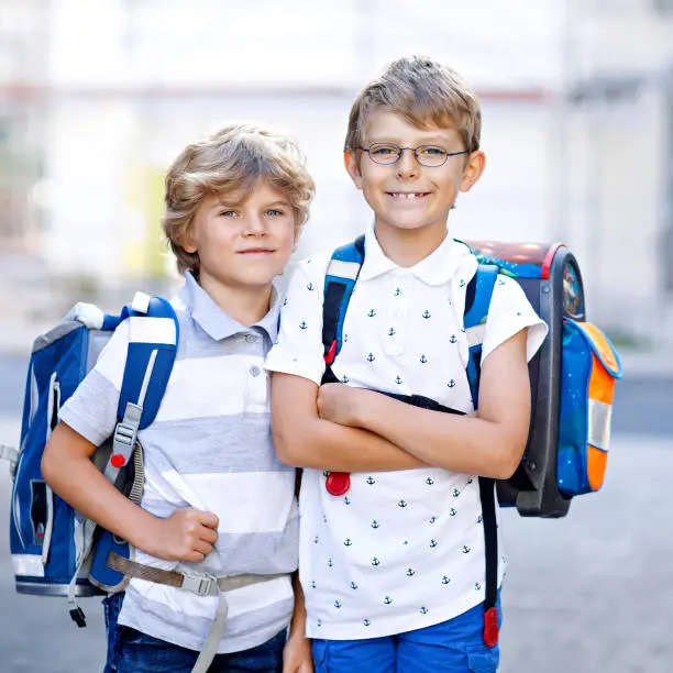 Two little kid boys with backpack or satchel. Schoolkids on the way to school. Healthy adorable children, brothers and best friends outdoors on the street leaving home. Back to school. Happy siblings