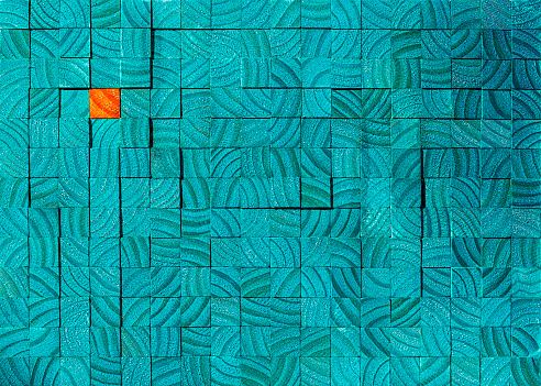 Many turquoise wooden blocks or profiles with the wood grain clearly visible, with one opposite color orange wooden block in contrast sitting amongst the turquoise ones. The color and wood grain creates a strong abstract pattern. Concept image relating to standing out from the crowd, against the grain, being different, individuality, singled out etc.