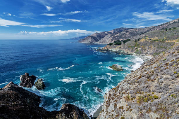 istock Shimmering reflective sunlight, blue skies, white clouds and waves splashing along the rugged Big Sur coastline, Big Creek Bridge viewed from a distance by Highway 1, on the California Central Coast. 1182653951