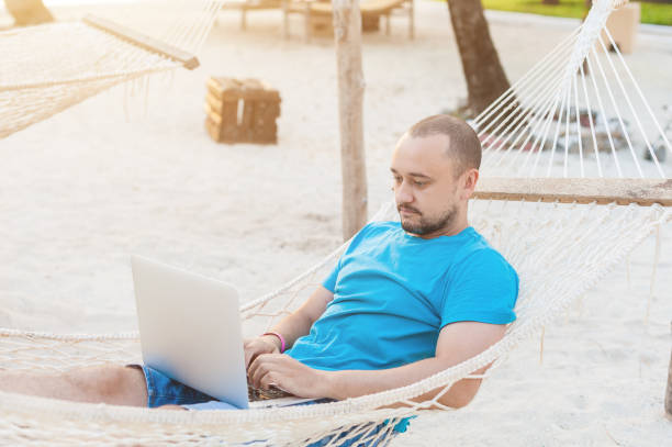 istock Man with a beard lying in a hammock with a laptop and looks at the screen. Work on the beach while traveling. The concept of remote work. 1182654847