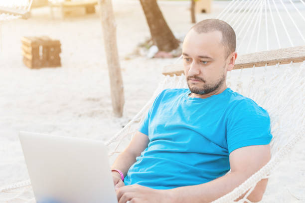 istock Man with a beard lying in a hammock with a laptop and looks at the screen. Work on the beach while traveling. The concept of remote work. 1182654914