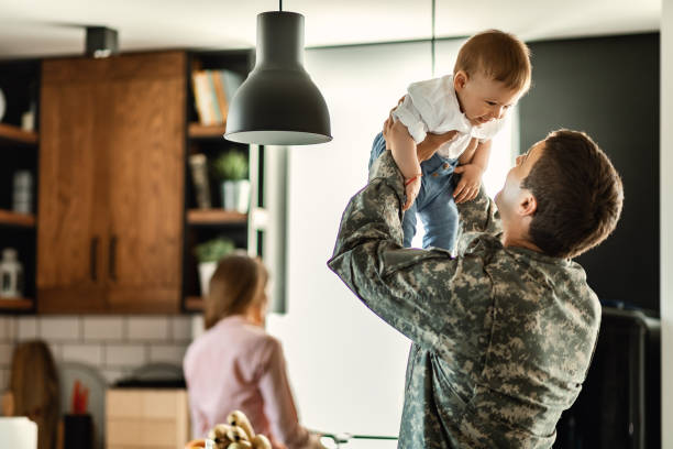Playful military man having fun with his small son at home. Happy baby boy having fun with his military dad at home. Mother is in the background. military lifestyle stock pictures, royalty-free photos & images