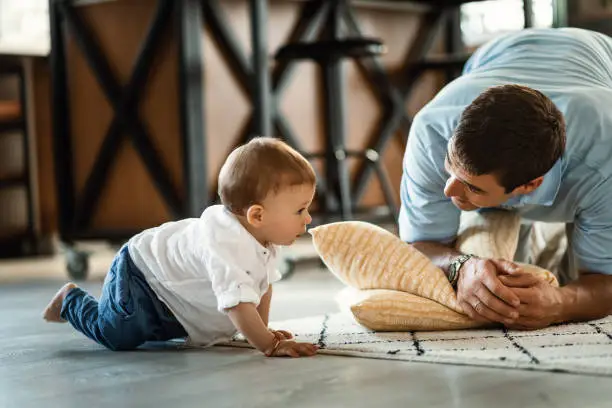 Happy father talking to his small son who is crawling on the floor in the living room.