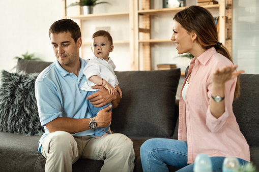 Married couple having an argument at home. Man is holding their small son, while woman is feeling angry and screaming at him.