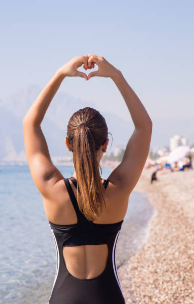 giovane donna sportiva fa la cornice del dito a forma di cuore sulla spiaggia - rear view women back back of head foto e immagini stock