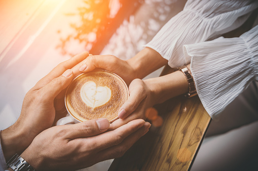 Valentine day, Close up of a young lovers hand holding a heart shaped coffee cup on a wooden table in the coffee shop. Warm Tone. Couple wedding.