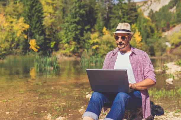 freelancer moderno usando portátil mientras se sienta en la roca en la costa del lago. espacio para copiar. - hipster people surfing the net internet fotografías e imágenes de stock