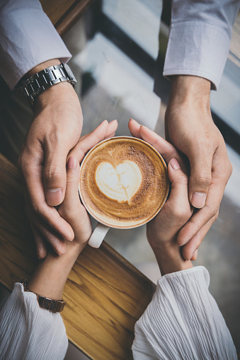 Valentine day, Top view of a young lovers hand holding a heart shaped coffee cup on a wooden table in the coffee shop. Warm Tone.