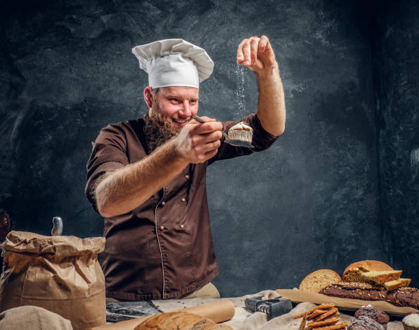 istock Cheerful bearded baker sprinkling some flour on his freshly made muffin next to a table 1182679398