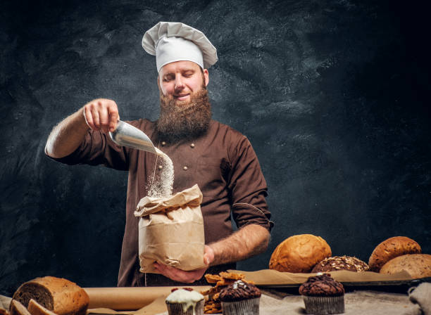 istock A bearded baker wearing a cook uniform sprinkling some flour in bag, standing next to a table, decorated with delicious bread loaves, baguettes 1182679418