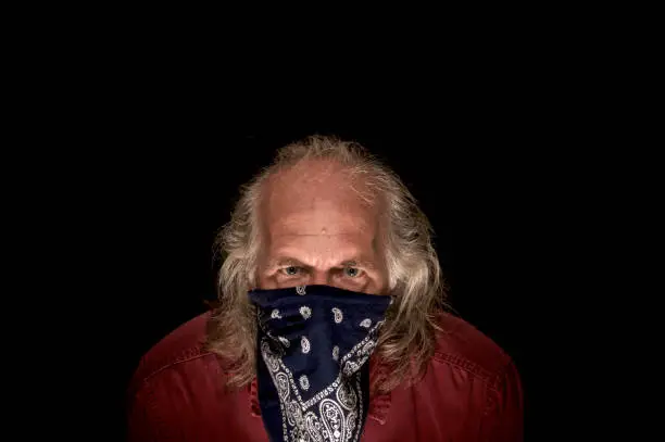 portrait of llong haired masked bandit wearing a blue bandana to cover the face and a red shirt looking intently at viewer with dark background.