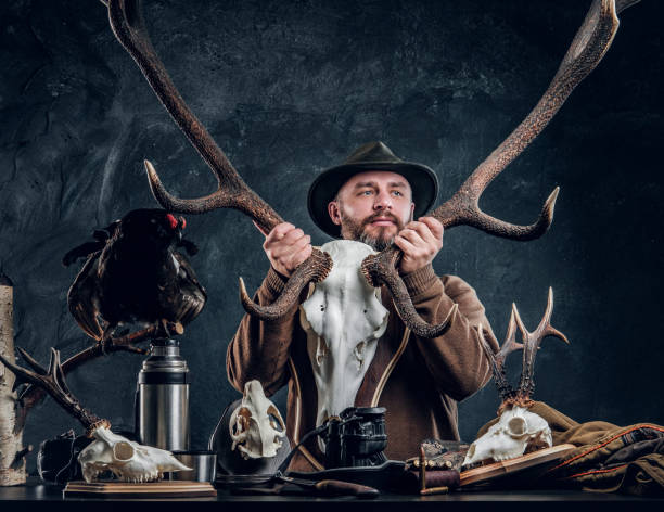 istock Mature bearded hunter in hat holding a big deer skull while standing next to a table with equipment and trophys. 1182682308